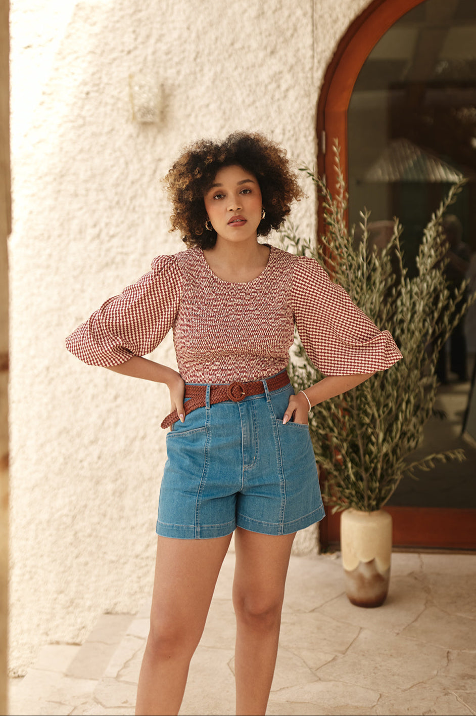 Woman in a burgundy shirred top and blue shorts standing in front of a textured wall with a plant.