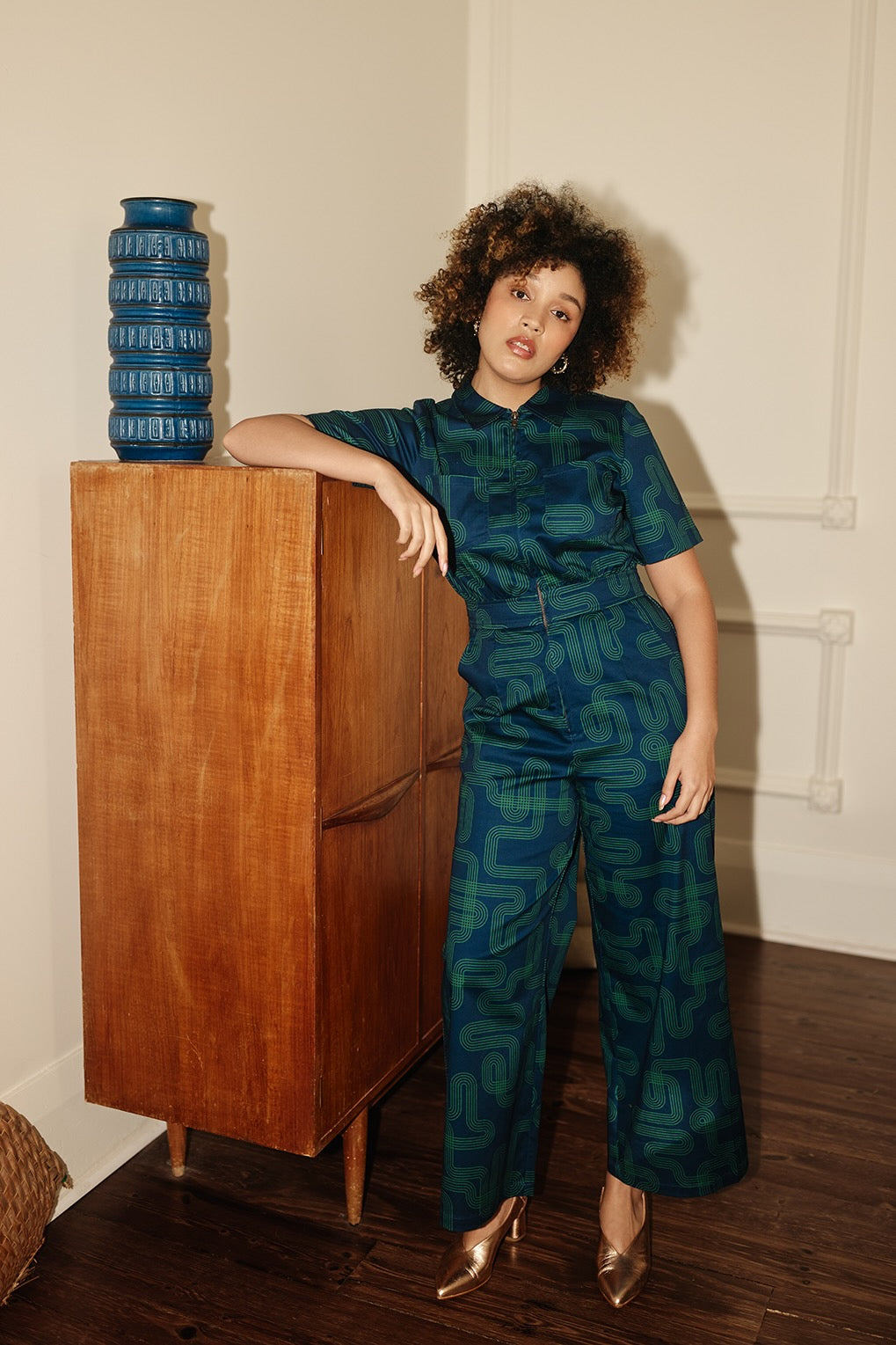 Woman standing next to wooden cabinet wearing Frock Me Out Labyrinth Freestyler Jumpsuit 