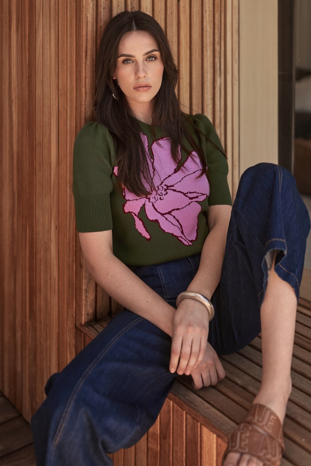 Woman wearing a green knit top with a lilac floral design and blue jeans sitting on a wooden bench.
