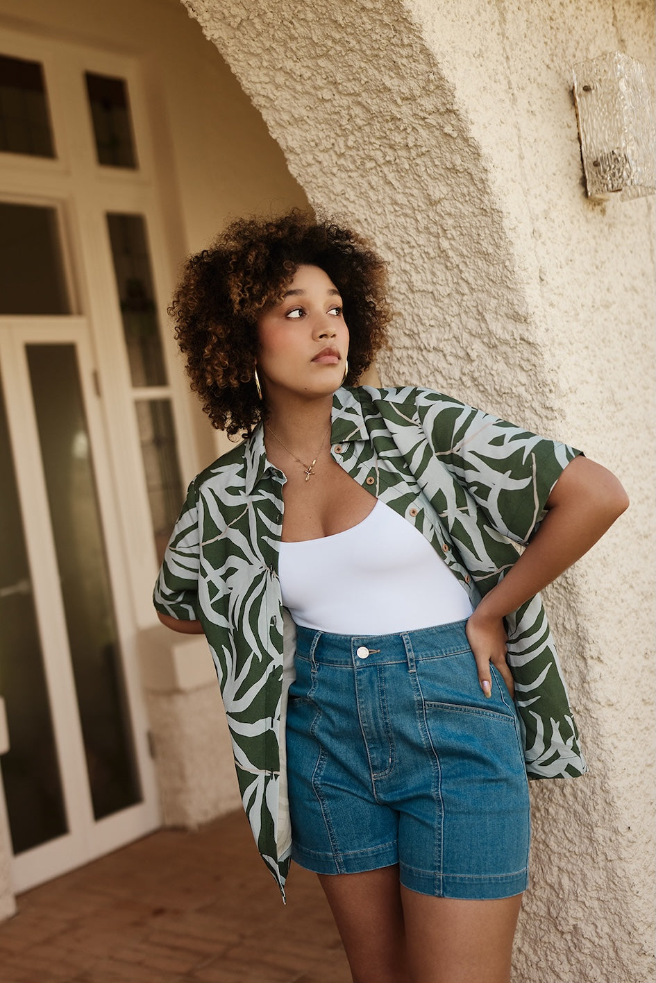 Woman wearing a green and white patterned shirt and blue shorts standing against a textured wall.